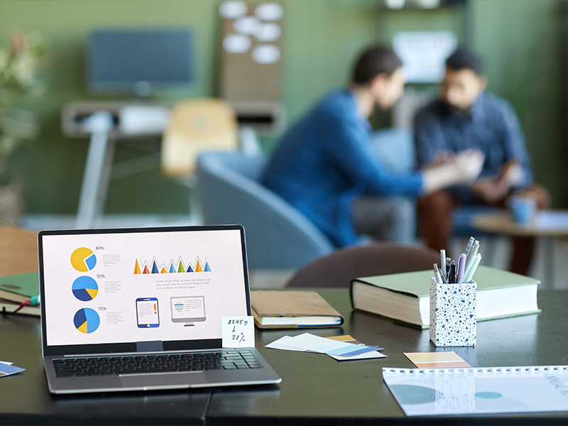 Laptop zeigt Diagramme und im Hintergrund sitzen zwei Personen